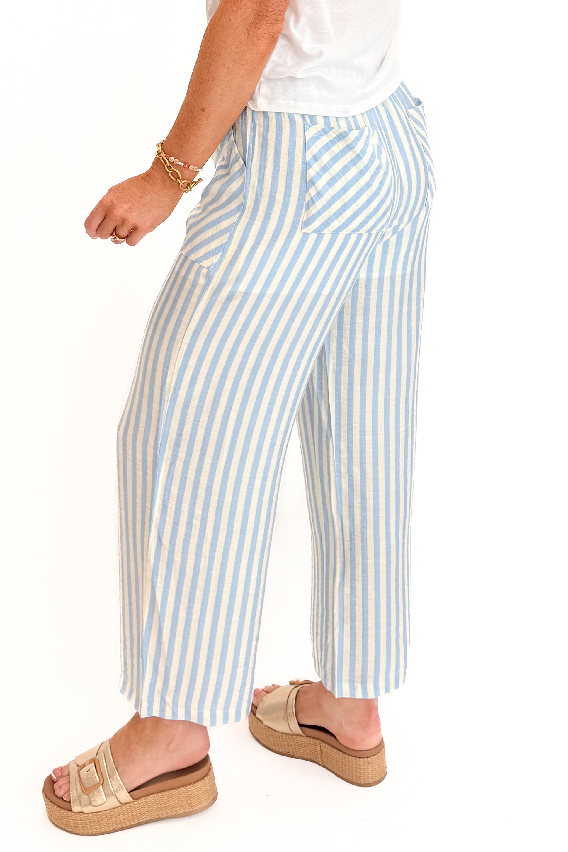 Side detail view of woman wearing Swagger Boutique’s Blue &amp; Cream Striped Pants with white top against white studio background.