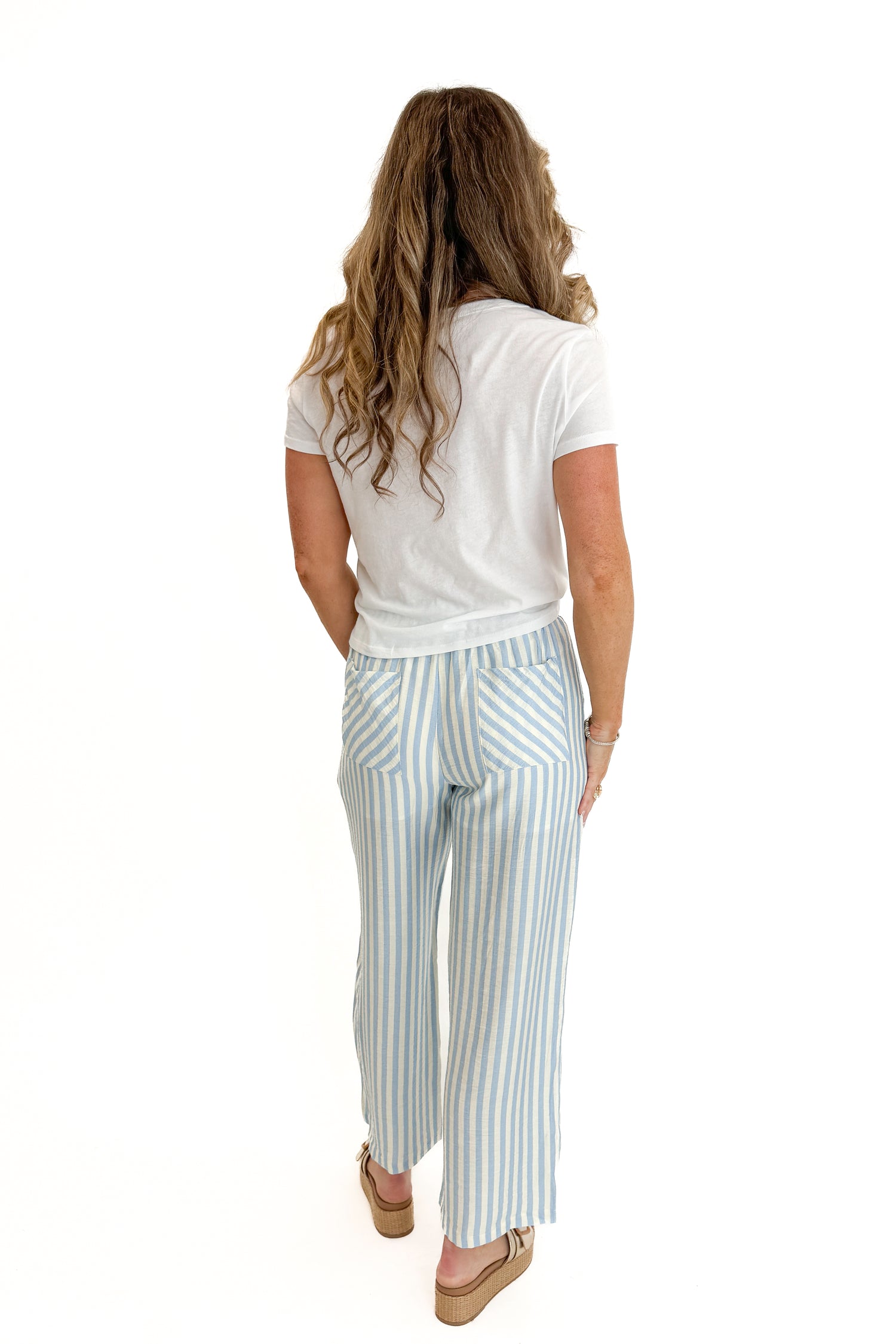 Back view of woman wearing Swagger Boutique’s Blue &amp; Cream Striped Pants with white top against white studio background.