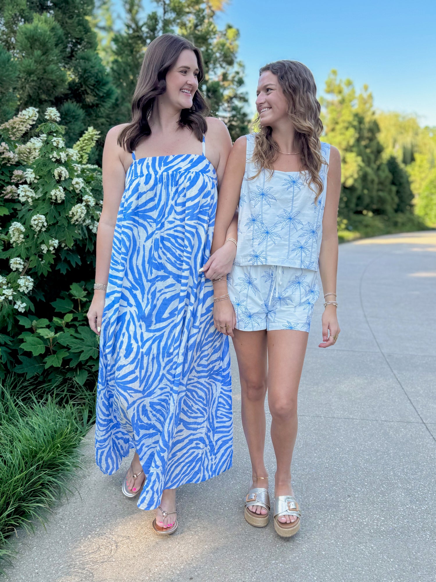 Front view of woman wearing Swagger Boutique’s Blue &amp; Cream Animal Print Maxi Dress by Entro and girl wearing White &amp; Blue Palm Embroidered Tank &amp; Shorts walking outside.