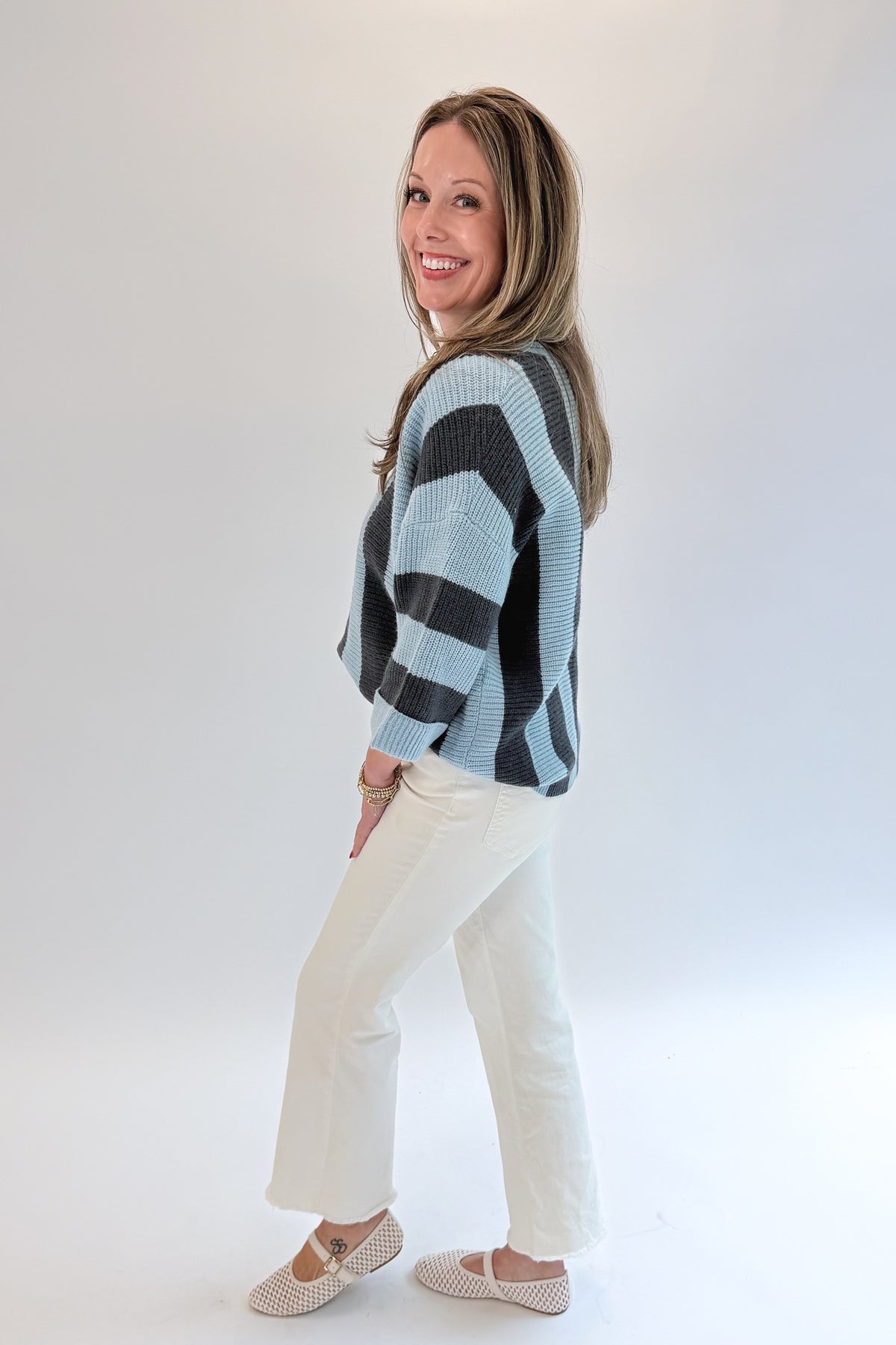 Side view of woman wearing Swagger Boutique’s Blue &amp; Grey Striped 3/4 Sleeve Sweater with jeans against white studio background.
