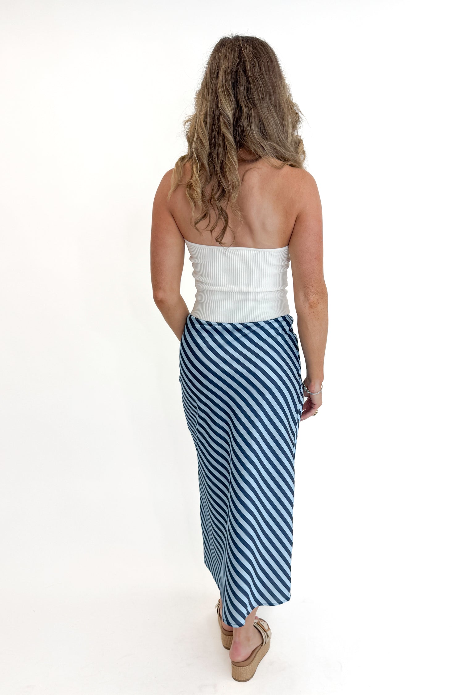 Back view of woman wearing Off White Ribbed Tube Top with Blue &amp; Navy Striped Satin Midi Skirt and sandals against white studio background.