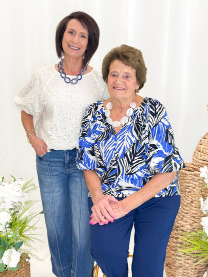 Front view of woman wearing Swagger Boutique’s Blue &amp; Navy Tropical Printed 3/4 Sleeve Top with blue pants against decorative background.