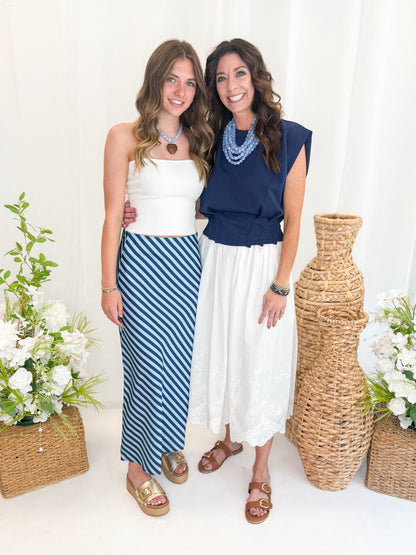 Front view of woman wearing Off White Ribbed Tube Top with Blue &amp; Navy Striped Satin Midi Skirt and sandals and woman wearing Navy Pleat Detail Tank with skirt against decorative background.