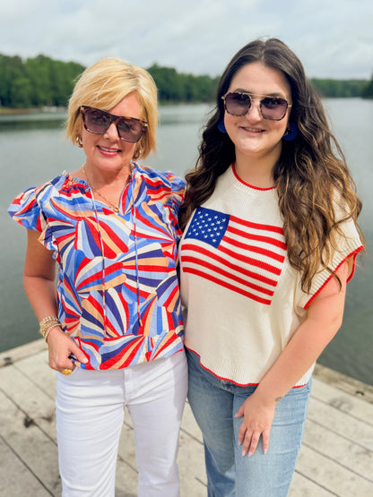 Front view of woman wearing Swagger Boutique’s Blue &amp; Red Printed Ruffle Sleeve Top by Skies Are Blue and woman wearing Flag sweater outside at the lake.