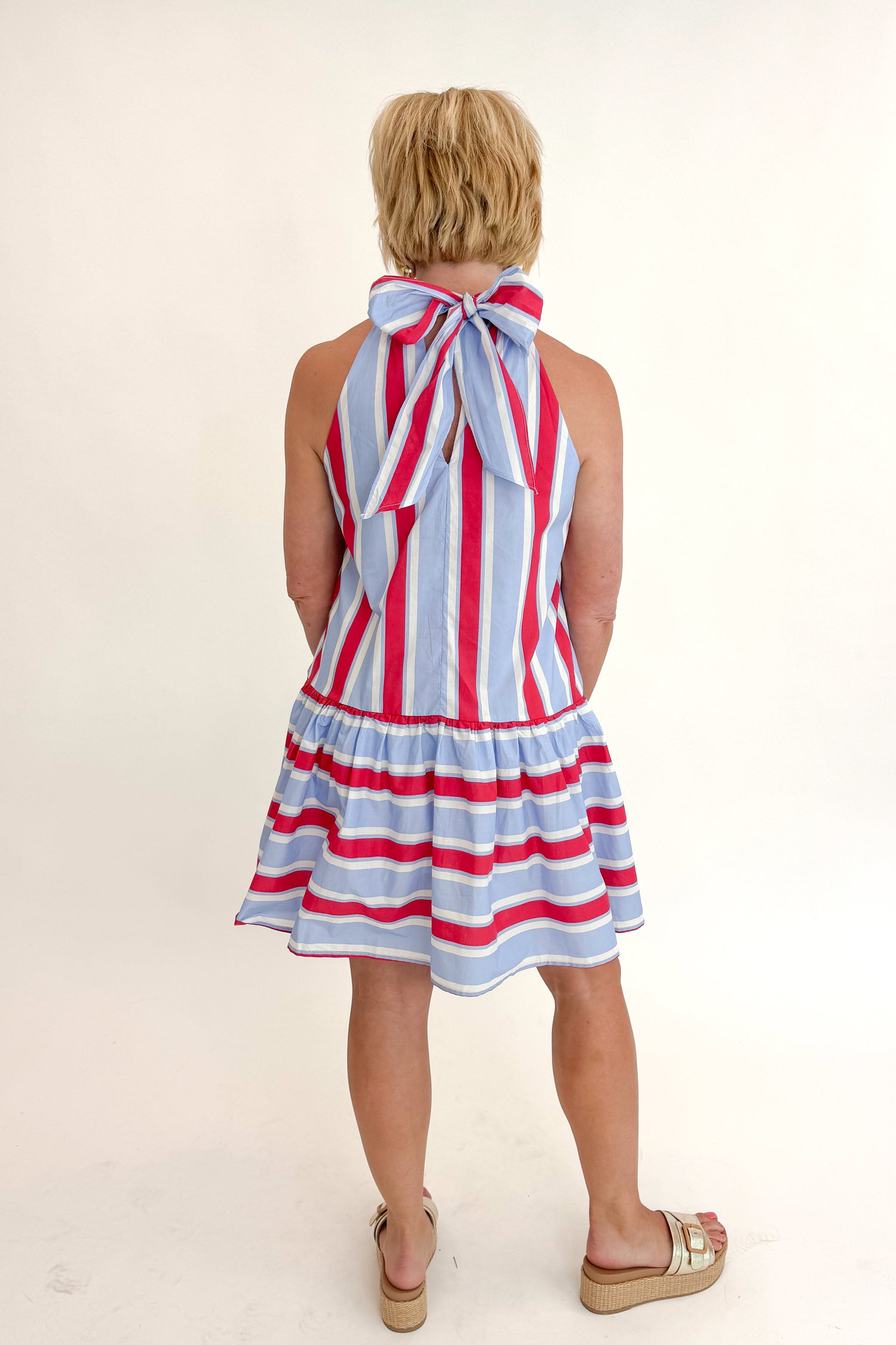 Back view of woman wearing Swagger Boutique’s Blue &amp; Red Striped Halter Neck Dress by Entro with sandals against white studio background.