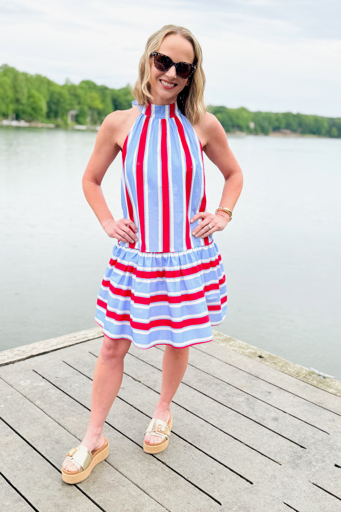 Front view of woman wearing Swagger Boutique’s Blue &amp; Red Striped Halter Neck Dress by Entro with Lulah Platform Sandals and sunglasses outside at the lake.