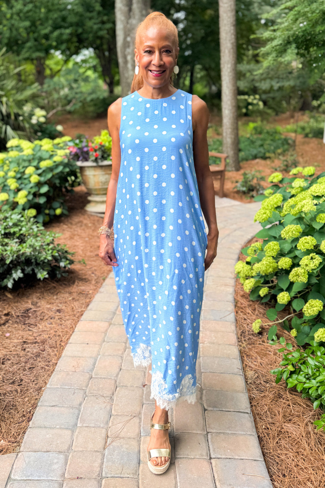 Front view of woman wearing Swagger Boutique’s Blue &amp; White Polka Dot Lace Trim Maxi Dress by Entro with wedges walking outside on a path.