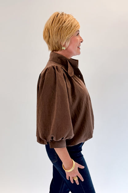 Side view of woman wearing Swagger Boutique’s Brown Bubble Hem Top With Buttons by Entro with jeans against white studio background.