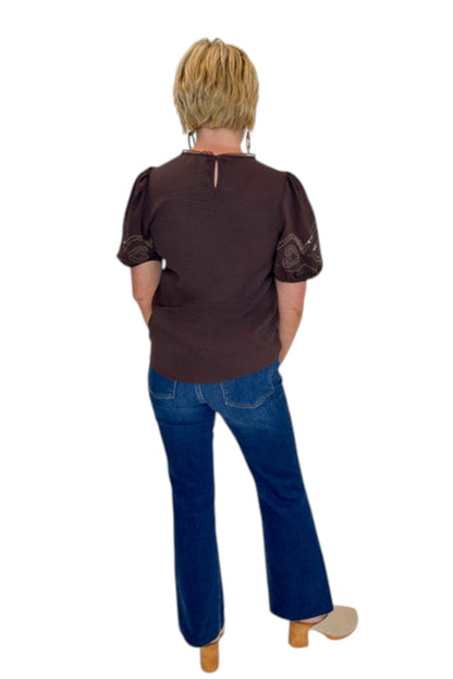 Back view of woman wearing Swagger Boutique’s Brown Embroidered Short Sleeve Top with jeans against white studio background.