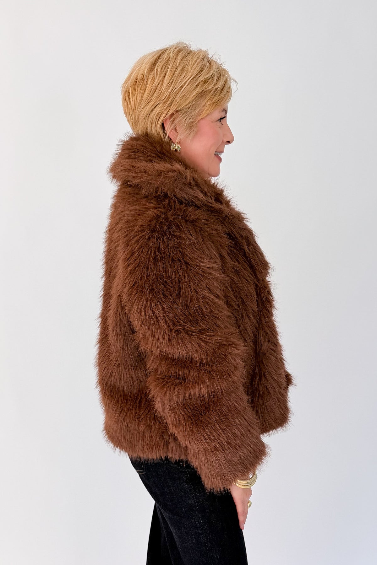 Side view of woman wearing Swagger Boutique’s Brown Faux Fur Jacket with jeans against white studio background.