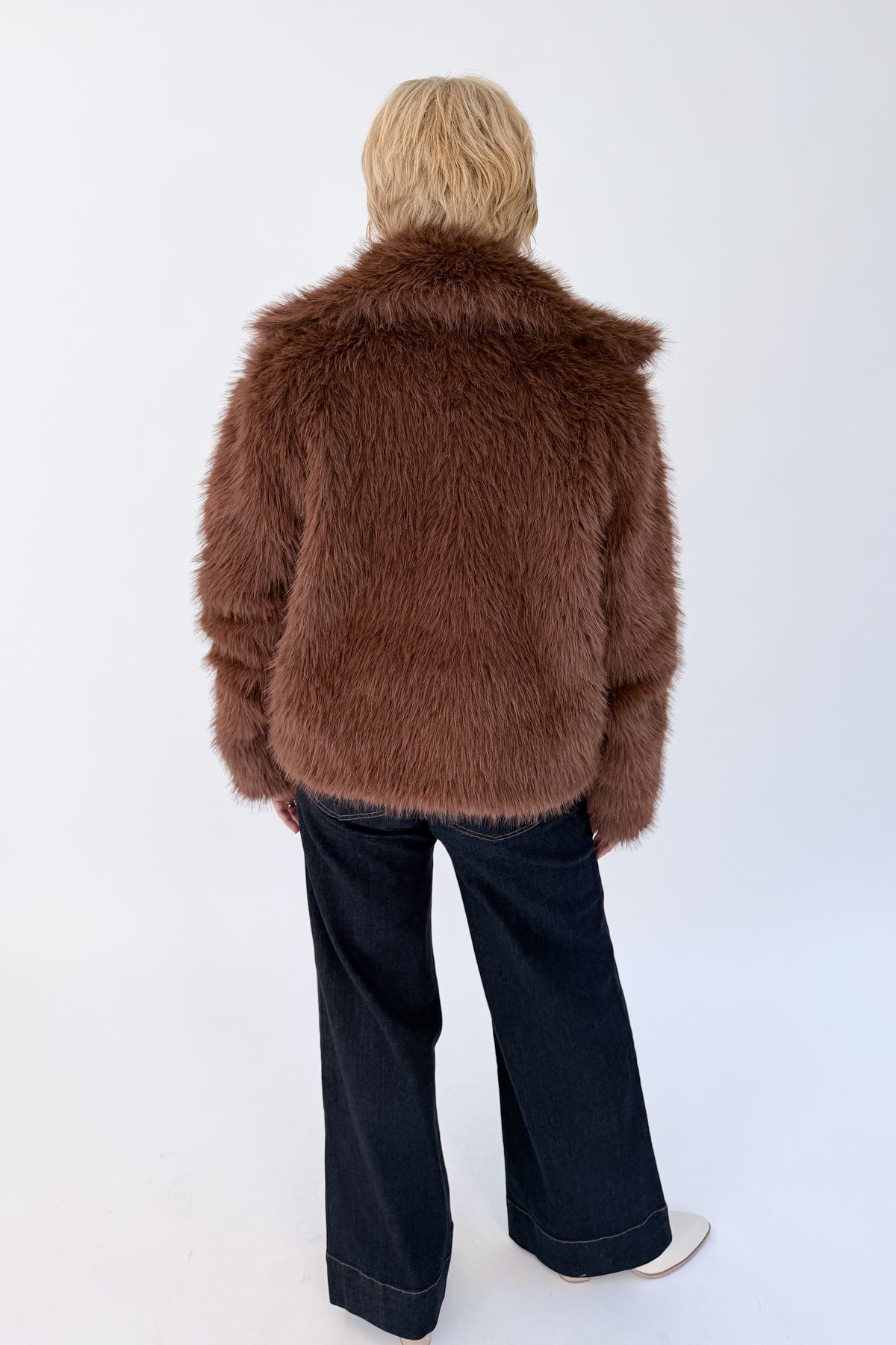 Back view of woman wearing Swagger Boutique’s Brown Faux Fur Jacket with jeans against white studio background.
