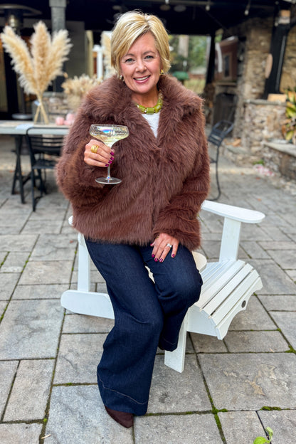 Front view of woman wearing Swagger Boutique’s Brown Faux Fur Jacket with jeans sitting outside at a party.