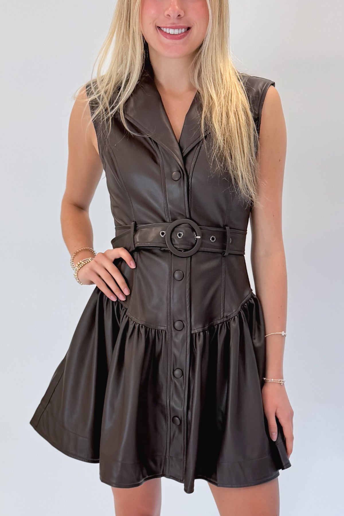 Front detail view of woman wearing Swagger Boutique’s Brown Faux Leather Mini Dress with heels against white studio background.