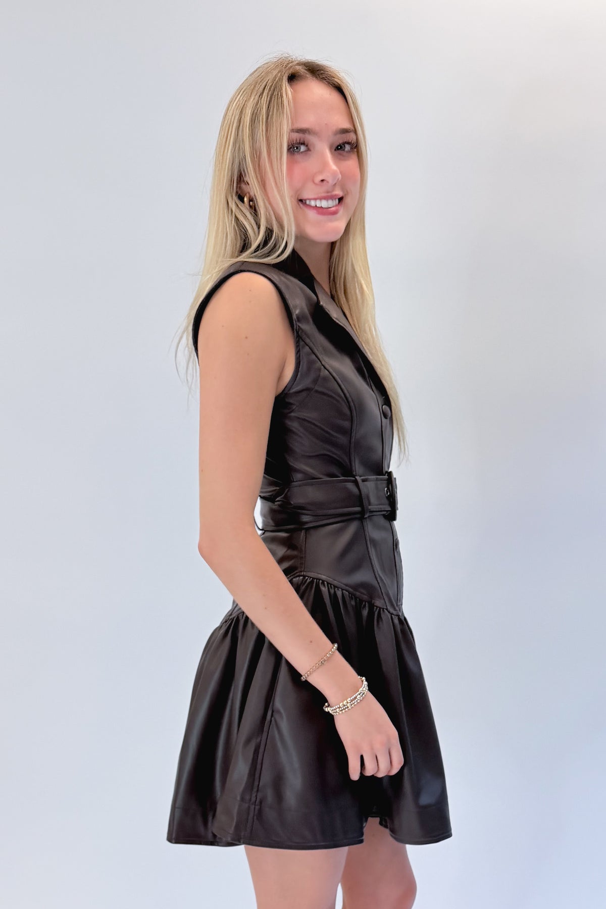 Side view of woman wearing Swagger Boutique’s Brown Faux Leather Mini Dress with heels against white studio background.