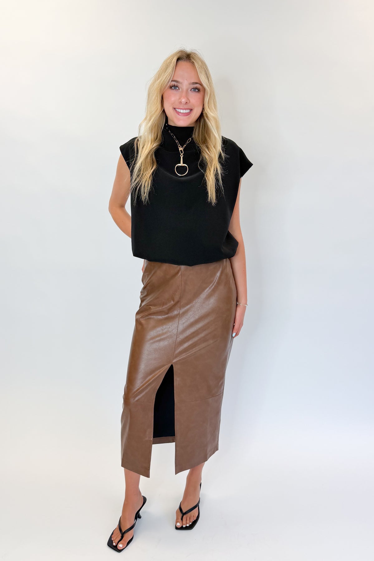Front view of woman wearing Swagger Boutique’s Brown Faux Leather Slit Midi Skirt with black top against white studio background.