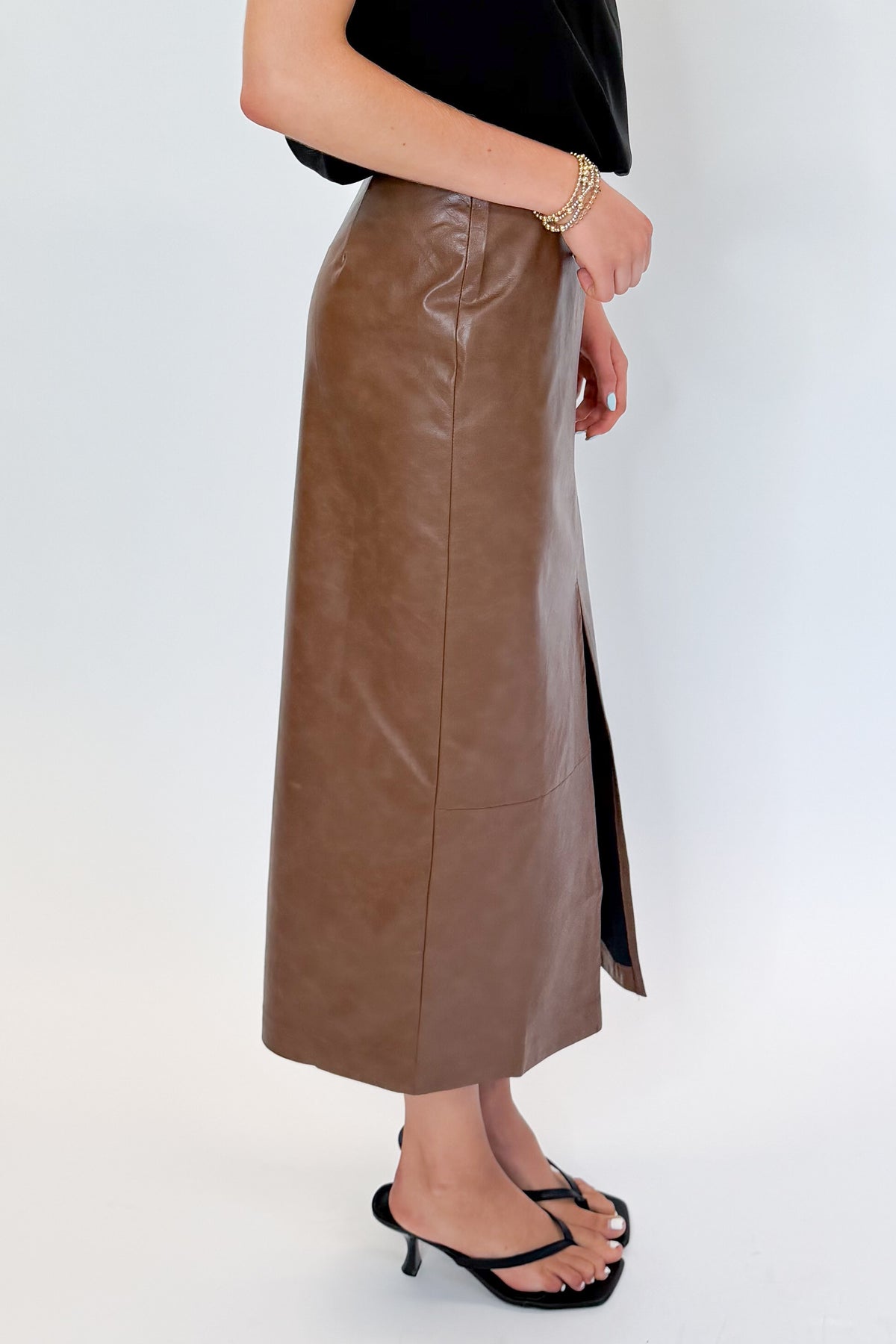 Side view of woman wearing Swagger Boutique’s Brown Faux Leather Slit Midi Skirt with black top against white studio background.