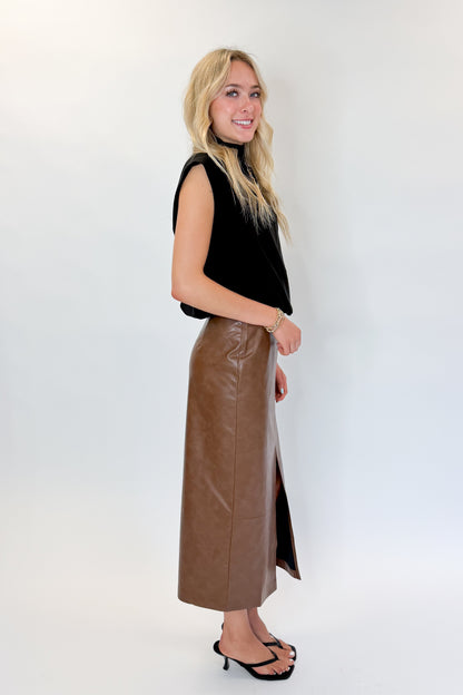 Side view of woman wearing Swagger Boutique’s Brown Faux Leather Slit Midi Skirt with black top against white studio background.