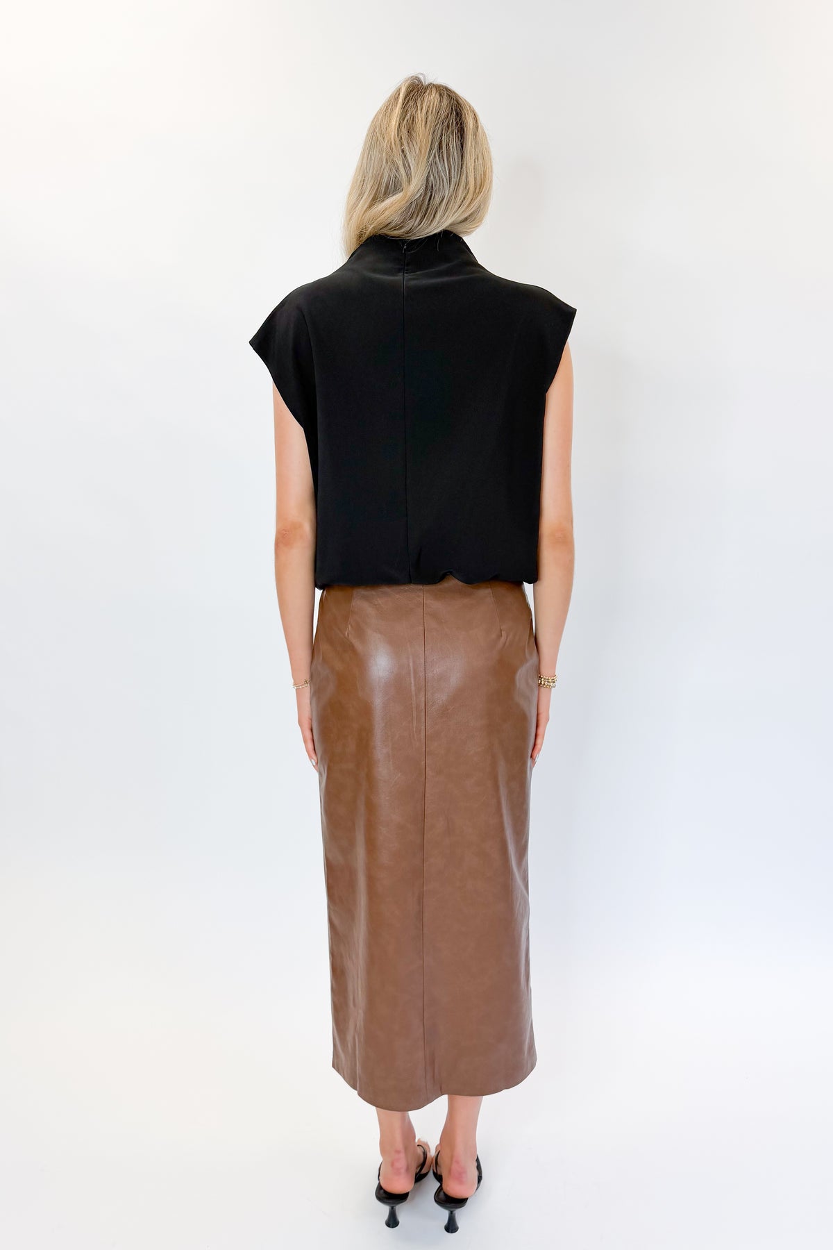 Back view of woman wearing Swagger Boutique’s Brown Faux Leather Slit Midi Skirt with black top against white studio background.