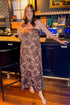 Front view of woman wearing Swagger Boutique’s Brown Floral Strapless Maxi Dress with heels standing at a restaurant bar area.