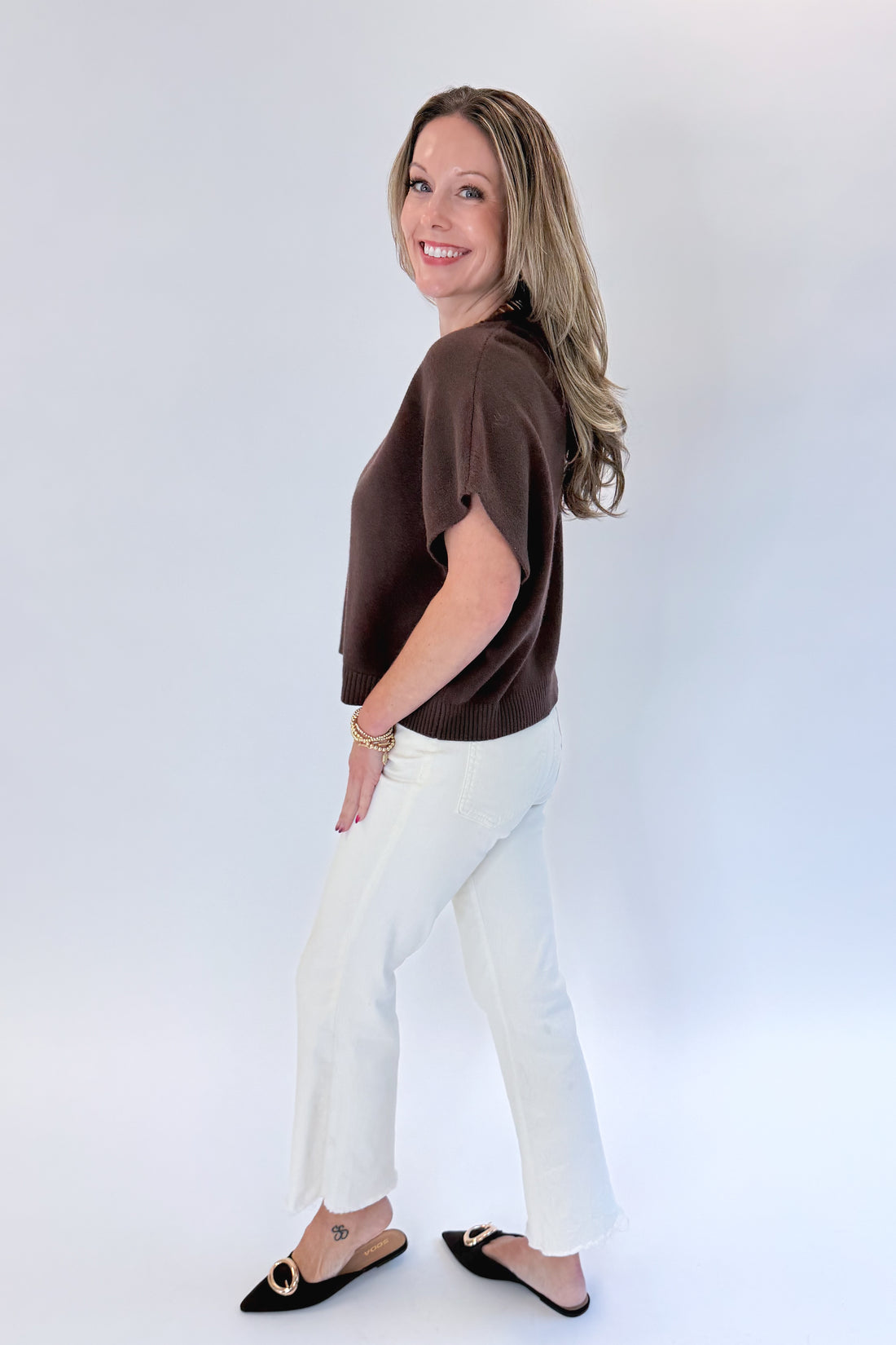 Side view of woman wearing Swagger Boutique’s Brown Light Weight Short Sleeve Sweater with jeans against white studio background.