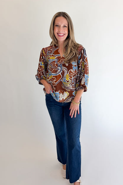 Front view of woman wearing Swagger Boutique’s Brown Paisley Print V-Neck Top with jeans against white studio background.