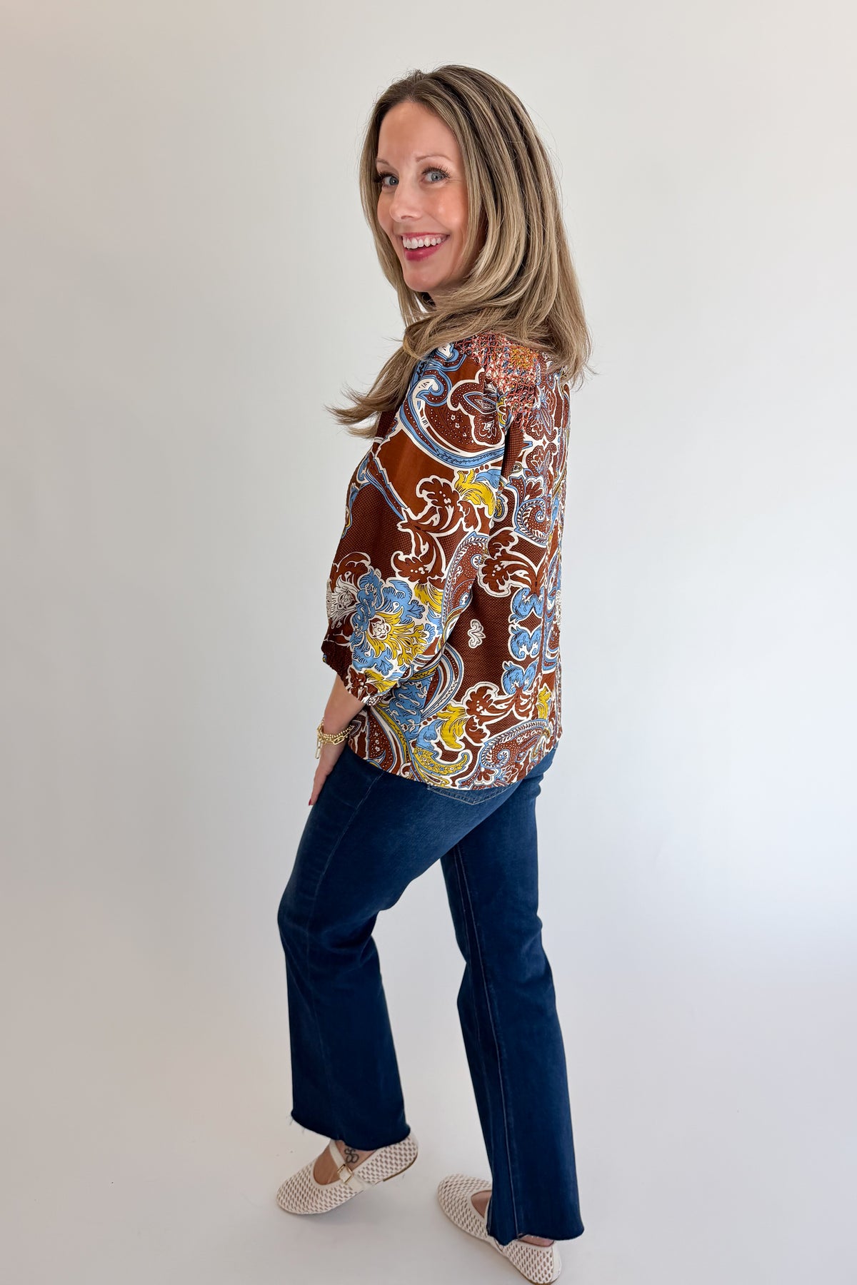 Side view of woman wearing Swagger Boutique’s Brown Paisley Print V-Neck Top with jeans against white studio background.