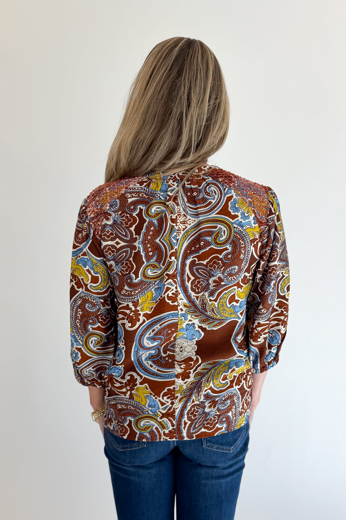 Back view of woman wearing Swagger Boutique’s Brown Paisley Print V-Neck Top with jeans against white studio background.