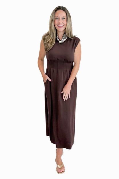 Front view of woman wearing Swagger Boutique’s Brown Pintuck Waist Midi Dress by Entro with sandals against white studio background.