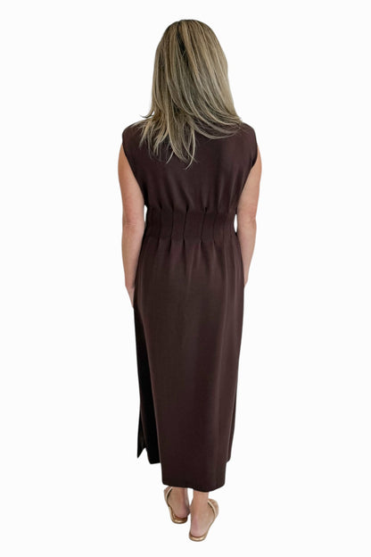 Back view of woman wearing Swagger Boutique’s Brown Pintuck Waist Midi Dress by Entro with sandals against white studio background.