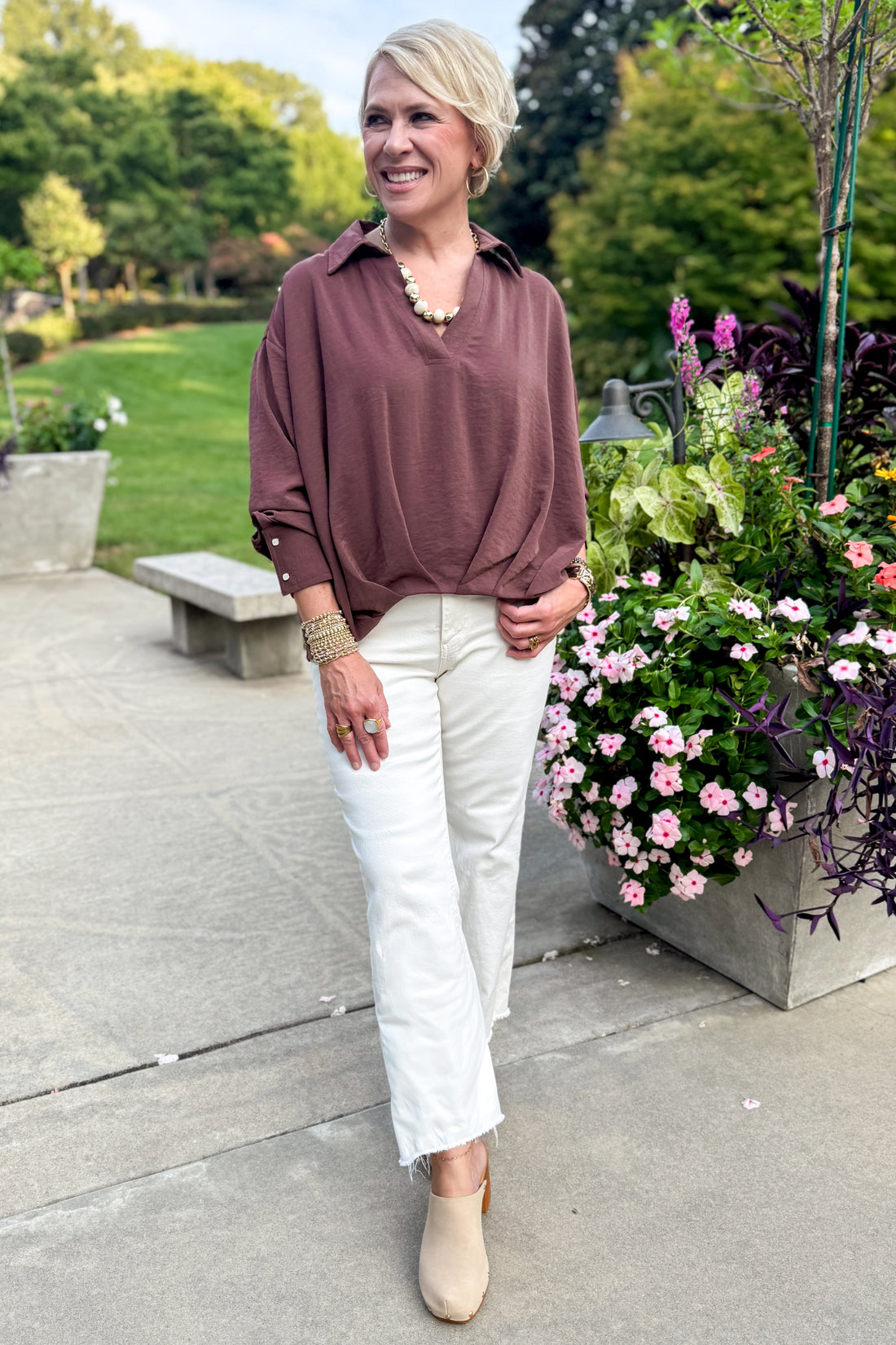 Front view of woman wearing Swagger Boutique’s Brown Pleated Long Sleeve Top by Entro with jeans standing outside by flowers. 