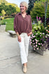 Front view of woman wearing Swagger Boutique’s Brown Pleated Long Sleeve Top by Entro with jeans standing outside by flowers. 