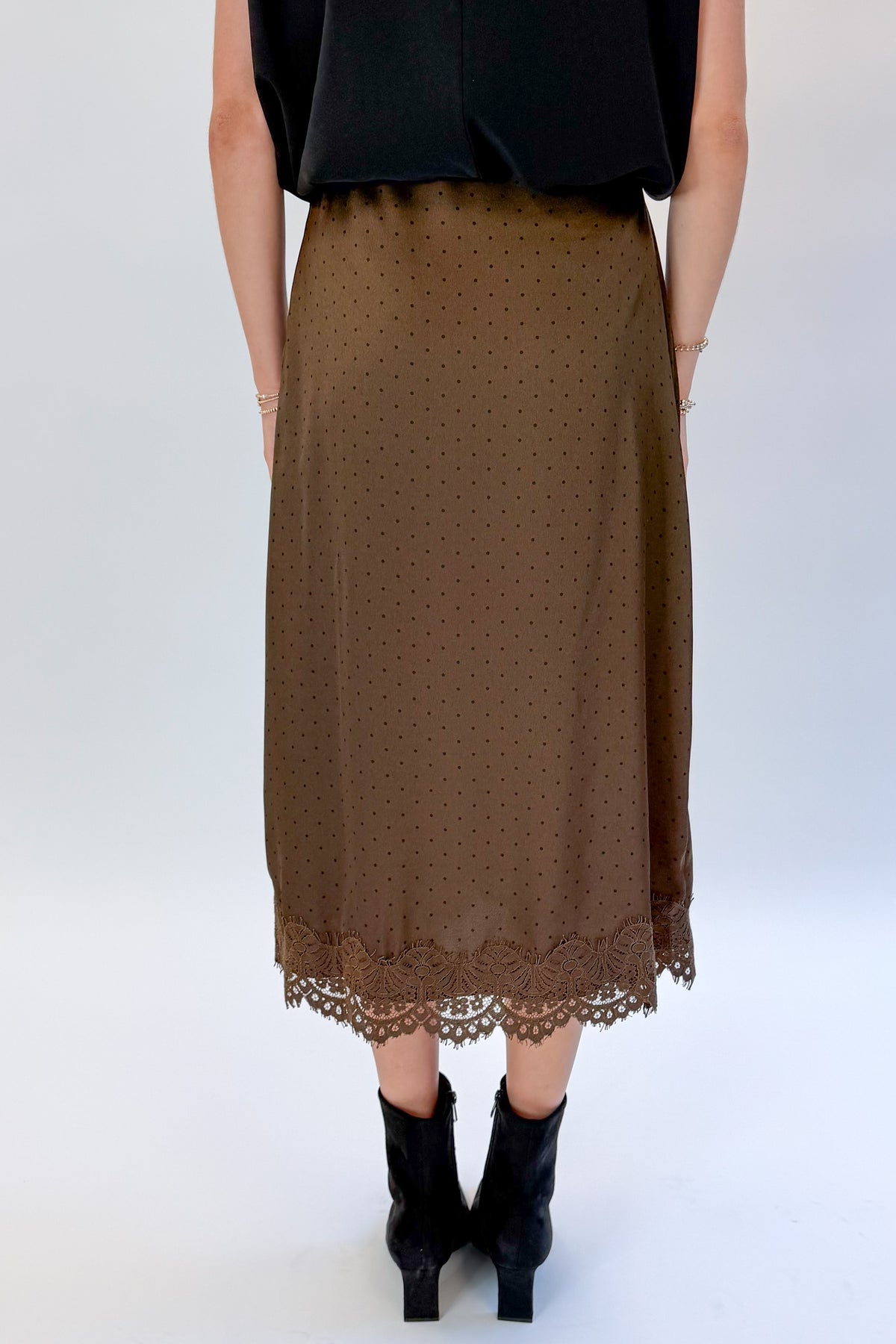 Back view of woman wearing Swagger Boutique’s Brown Polka Dot Lace Lined Satin Skirt by Entro with Black Satin Cowlneck Sleeveless Top against white studio background.