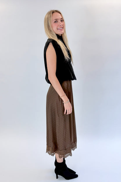 Side view of woman wearing Swagger Boutique’s Brown Polka Dot Lace Lined Satin Skirt by Entro with Black Satin Cowlneck Sleeveless Top against white studio background.