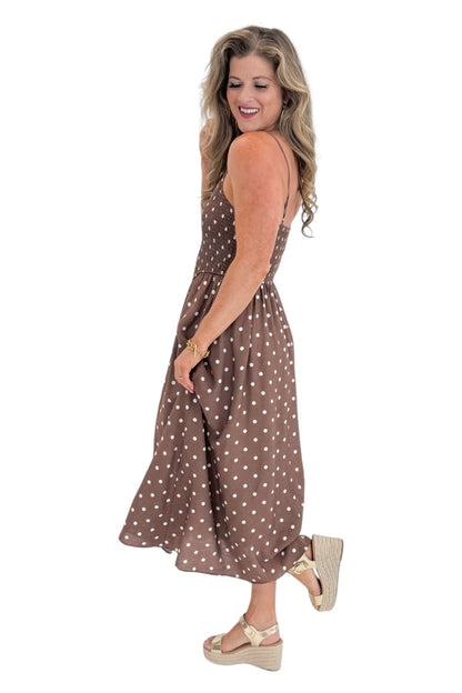 Side view of woman wearing Swagger Boutique’s Brown Polka Dot Smocked Midi Dress with sandals against white studio background.