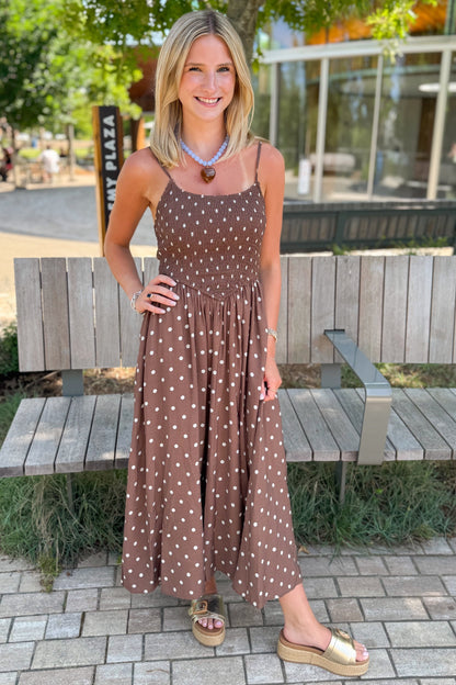 Front view of woman wearing Swagger Boutique’s Brown Polka Dot Smocked Midi Dress with sandals standing by a bench outside.