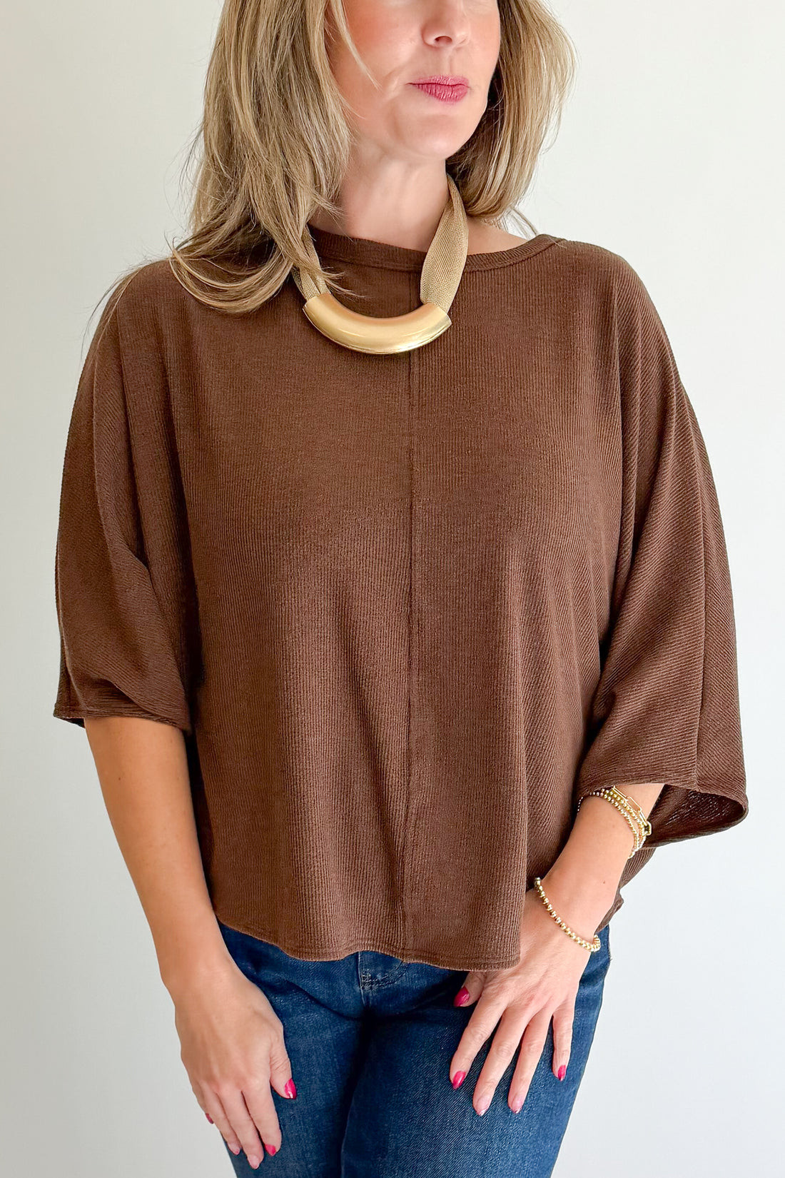 A woman wearing a brown short-sleeve knit top with three-quarter sleeves, paired with blue jeans and a chunky beige statement necklace, photographed against a plain light background.