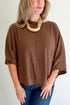 A woman wearing a brown short-sleeve knit top with three-quarter sleeves, paired with blue jeans and a chunky beige statement necklace, photographed against a plain light background.