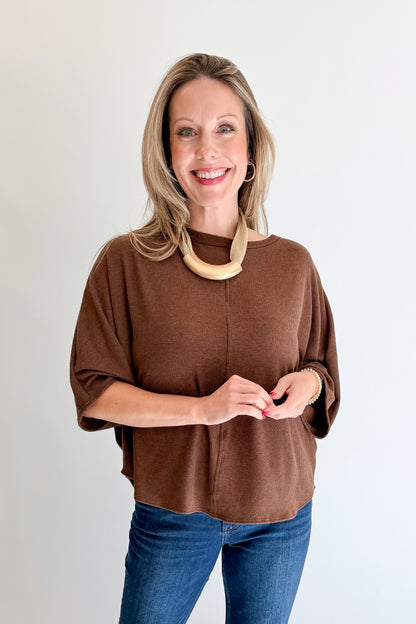 A smiling blonde woman wearing a brown short-sleeve knit top and blue jeans, standing against a plain light gray background.