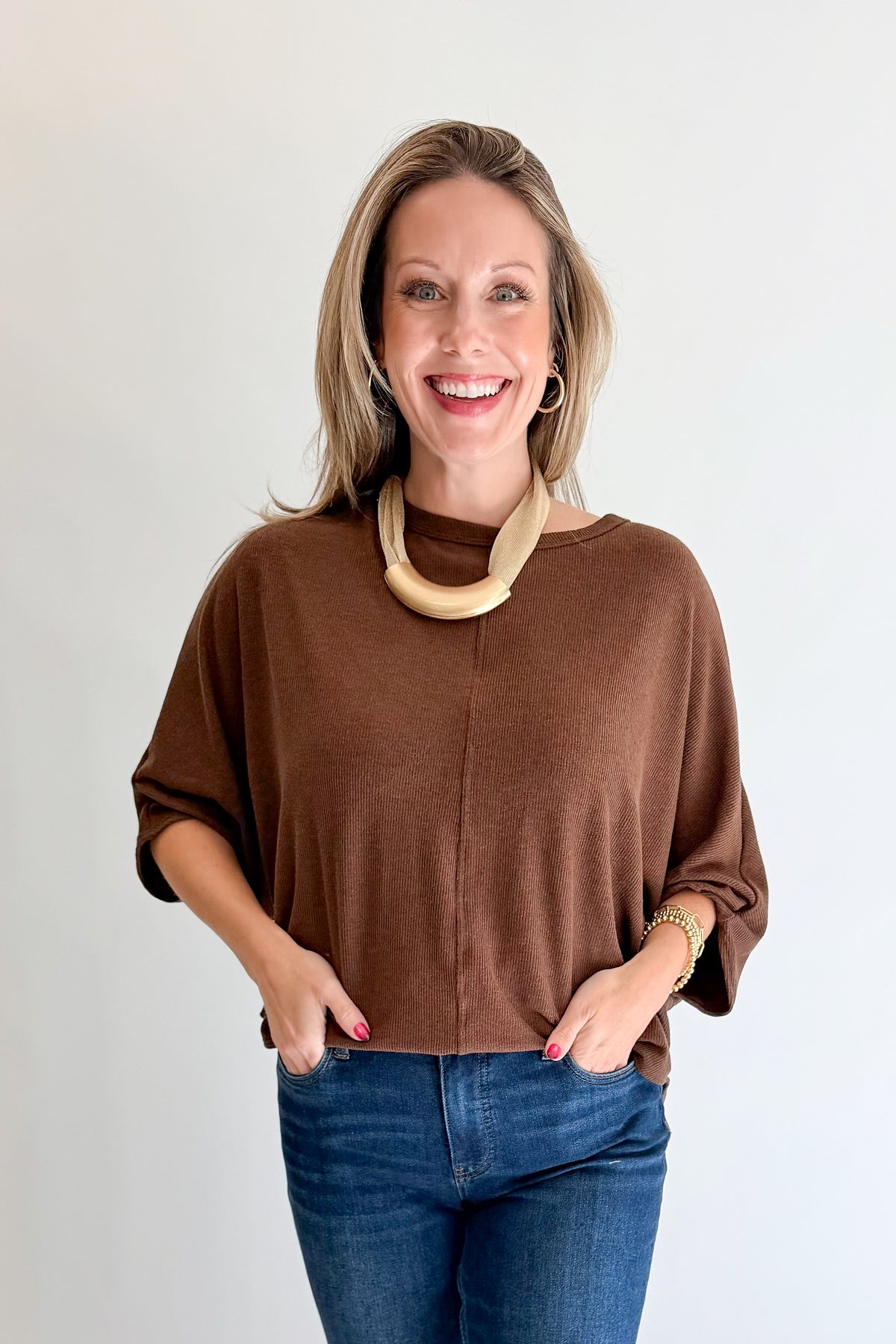 A smiling woman with blonde hair wearing a brown long-sleeve knit top and blue jeans, standing against a plain light gray background.