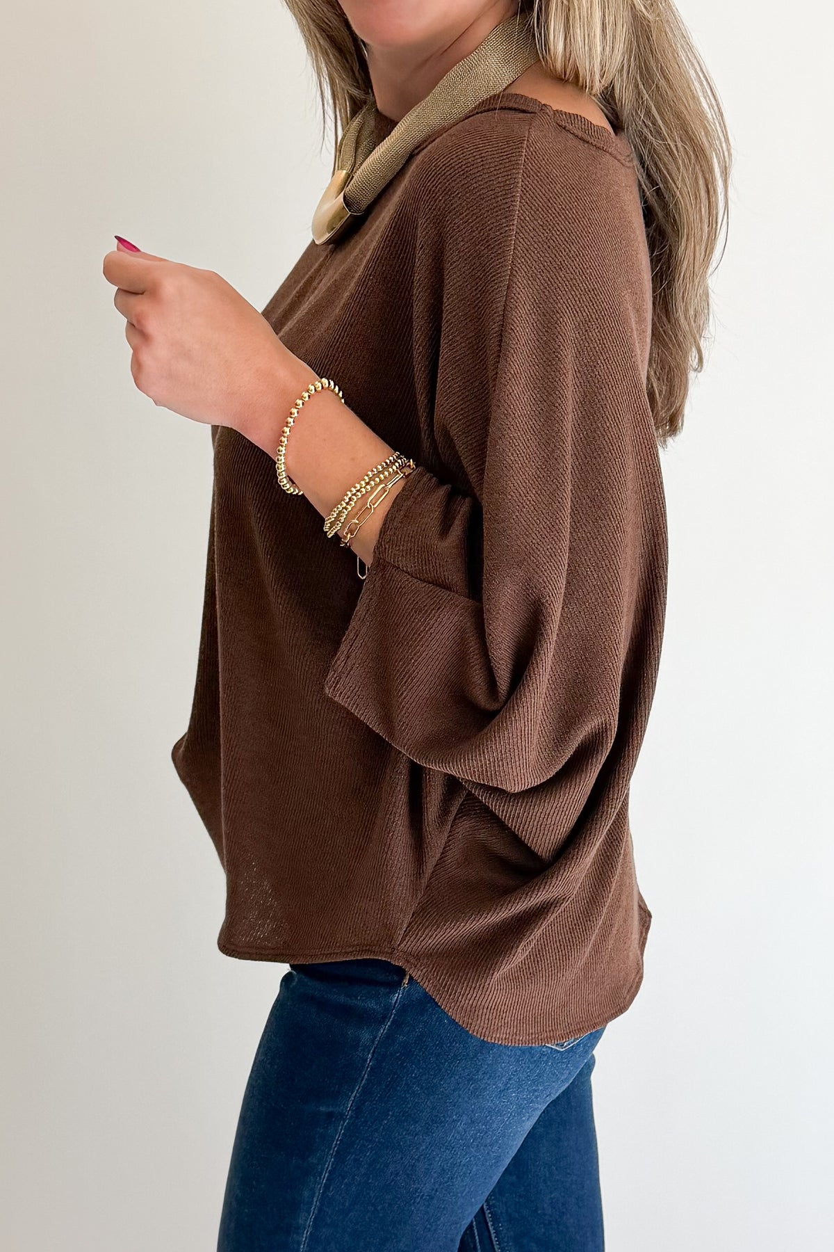 A woman wearing a brown knit long-sleeve top with a beige collar, paired with blue jeans, shown from the side against a plain light background.
