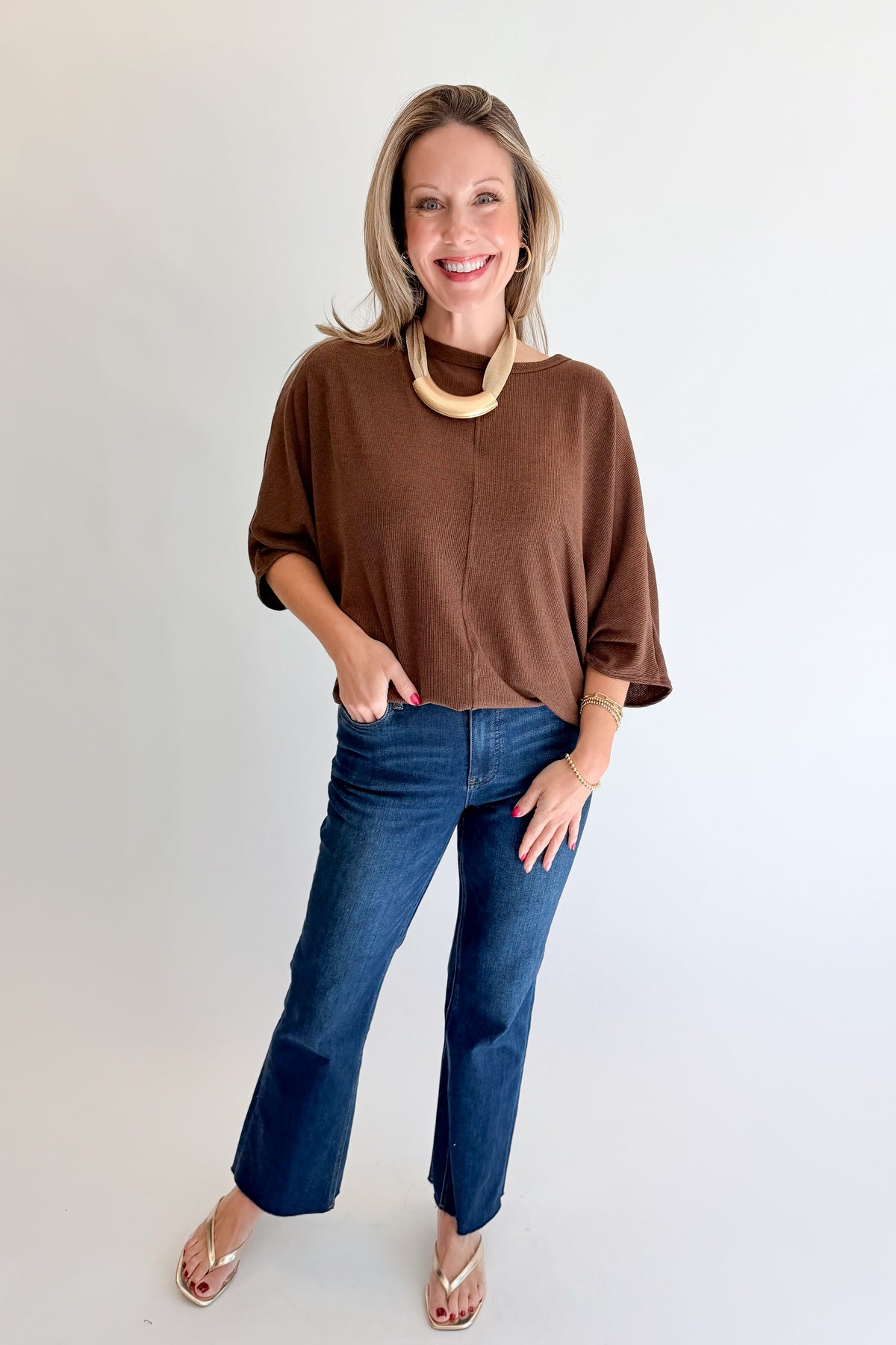 A woman modeling a brown short-sleeve knit top with blue jeans and a chunky beige necklace, standing against a plain white studio background.