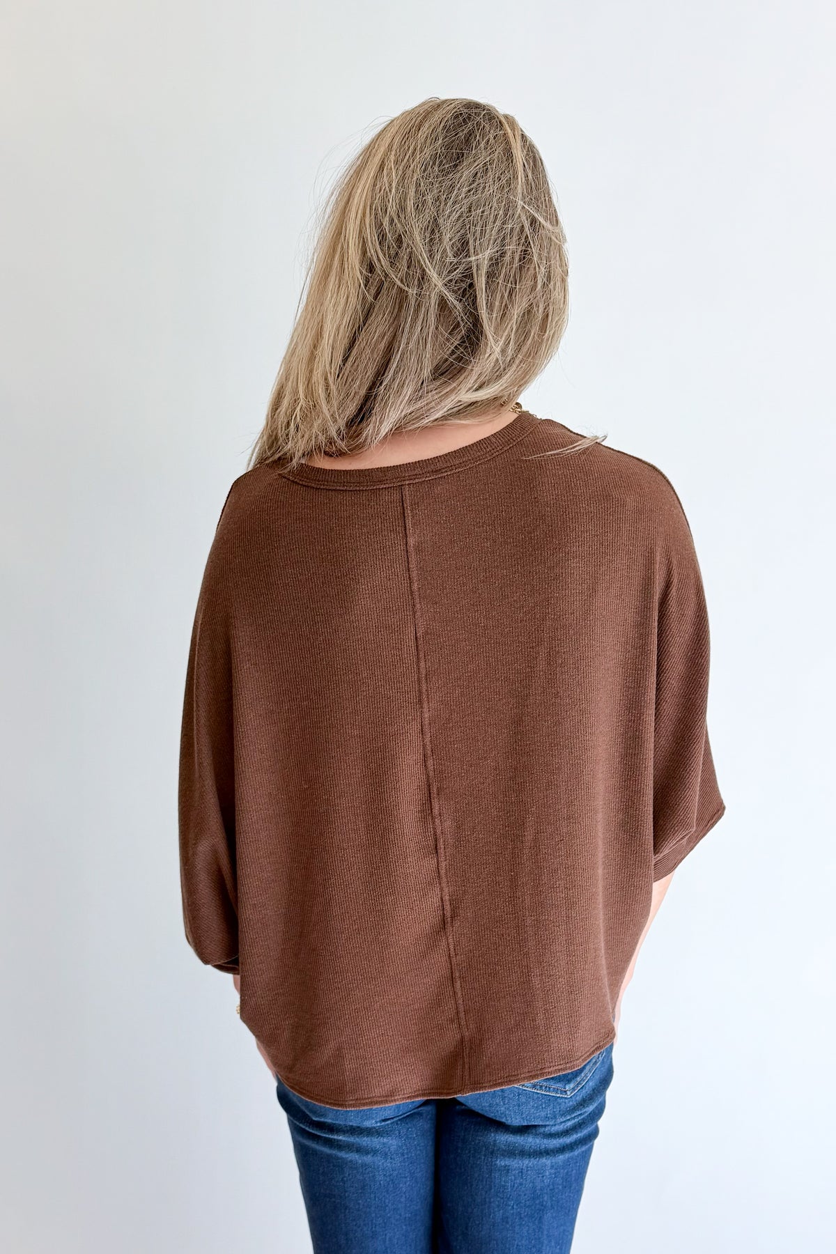 A woman viewed from behind wearing a loose-fitting brown short-sleeve top and blue jeans against a plain white background.
