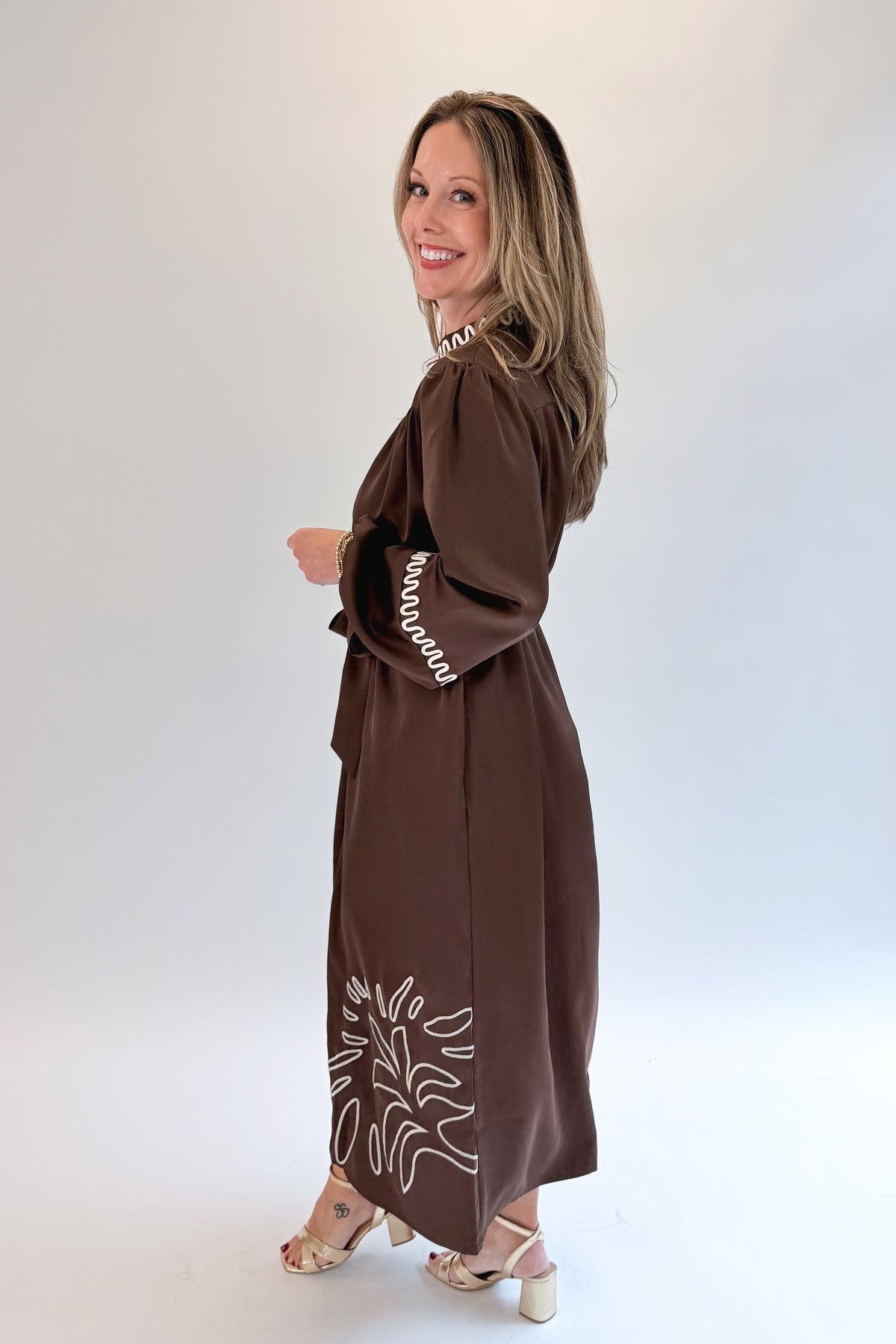 Side view of woman wearing Swagger Boutique’s Brown Satin Embroidered Midi Dress with heels against white studio background.