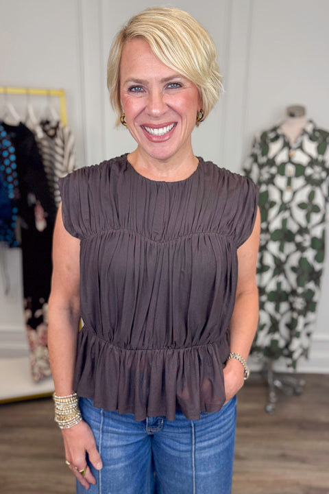 Front view of woman wearing Swagger Boutique’s Brown Shirred Peplum Top with jeans with clothing hanging in the background.