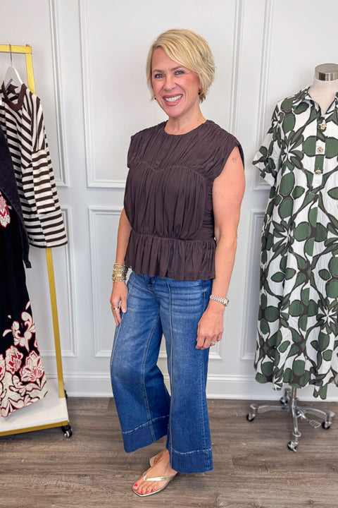 Side view of woman wearing Swagger Boutique’s Brown Shirred Peplum Top with jeans with clothing hanging in the background.