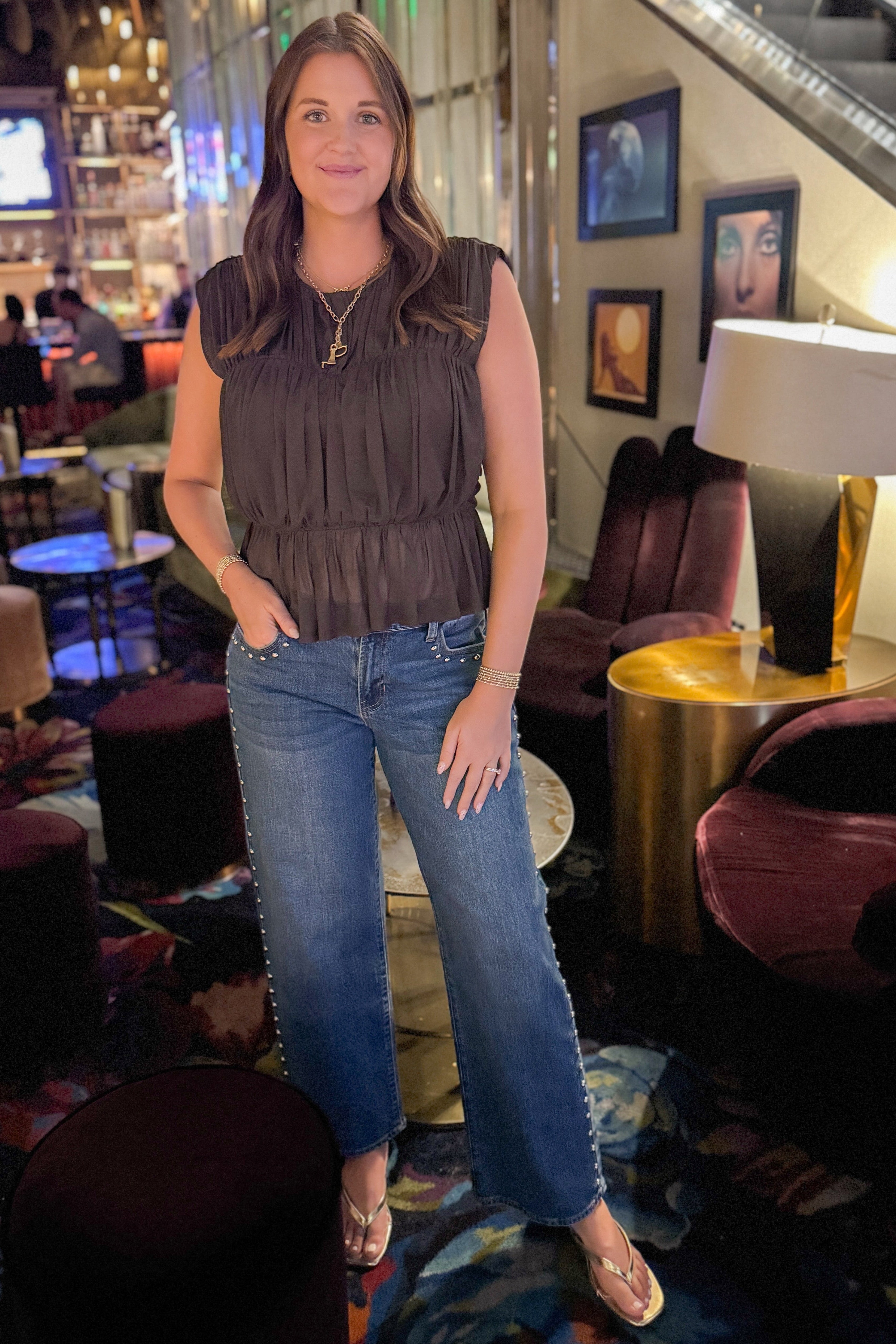 Front view of woman wearing Swagger Boutique’s Brown Shirred Peplum Top with jeans standing at a lounge.