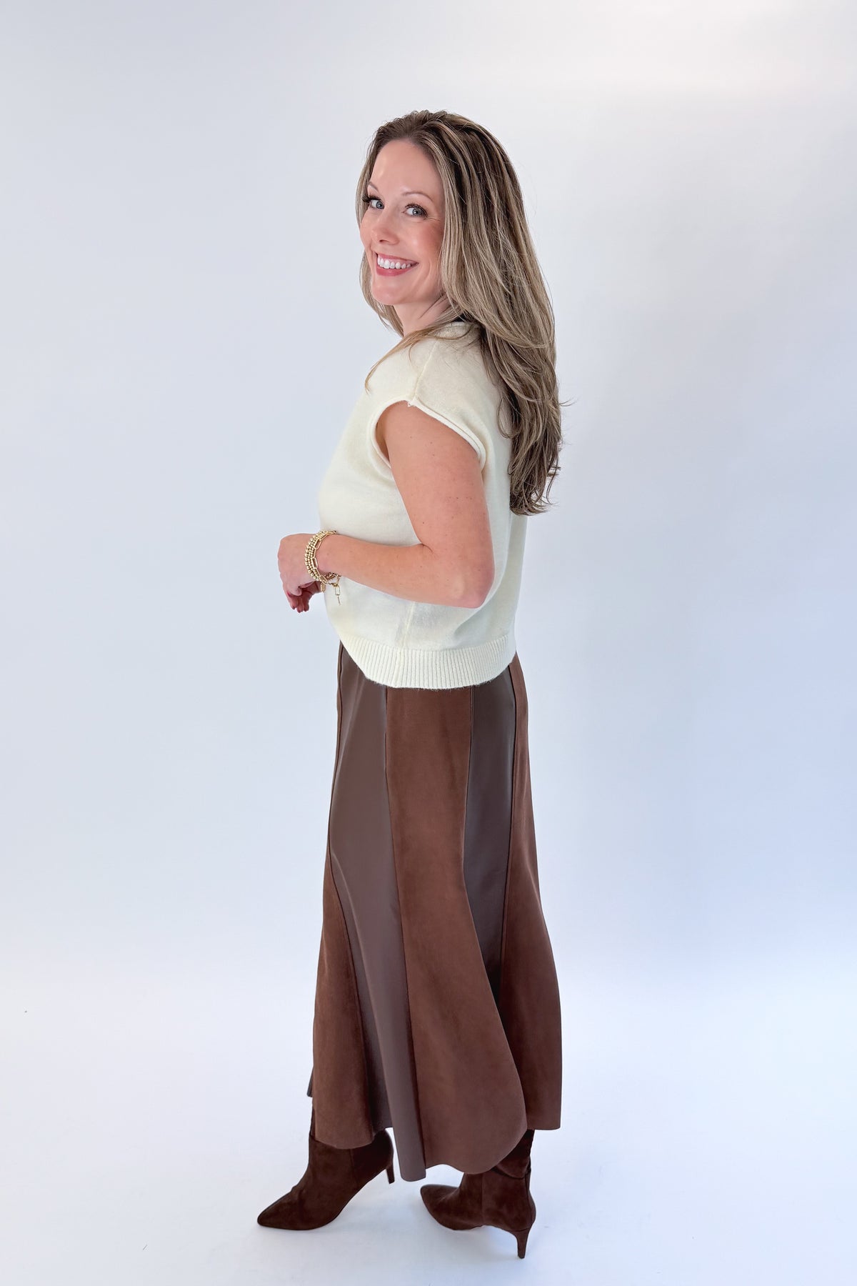 Side view of woman wearing Swagger Boutique’s Cream Sleeveless Button Up Sweater with Brown Suede &amp; Leather Midi Skirt by THML against white studio background.