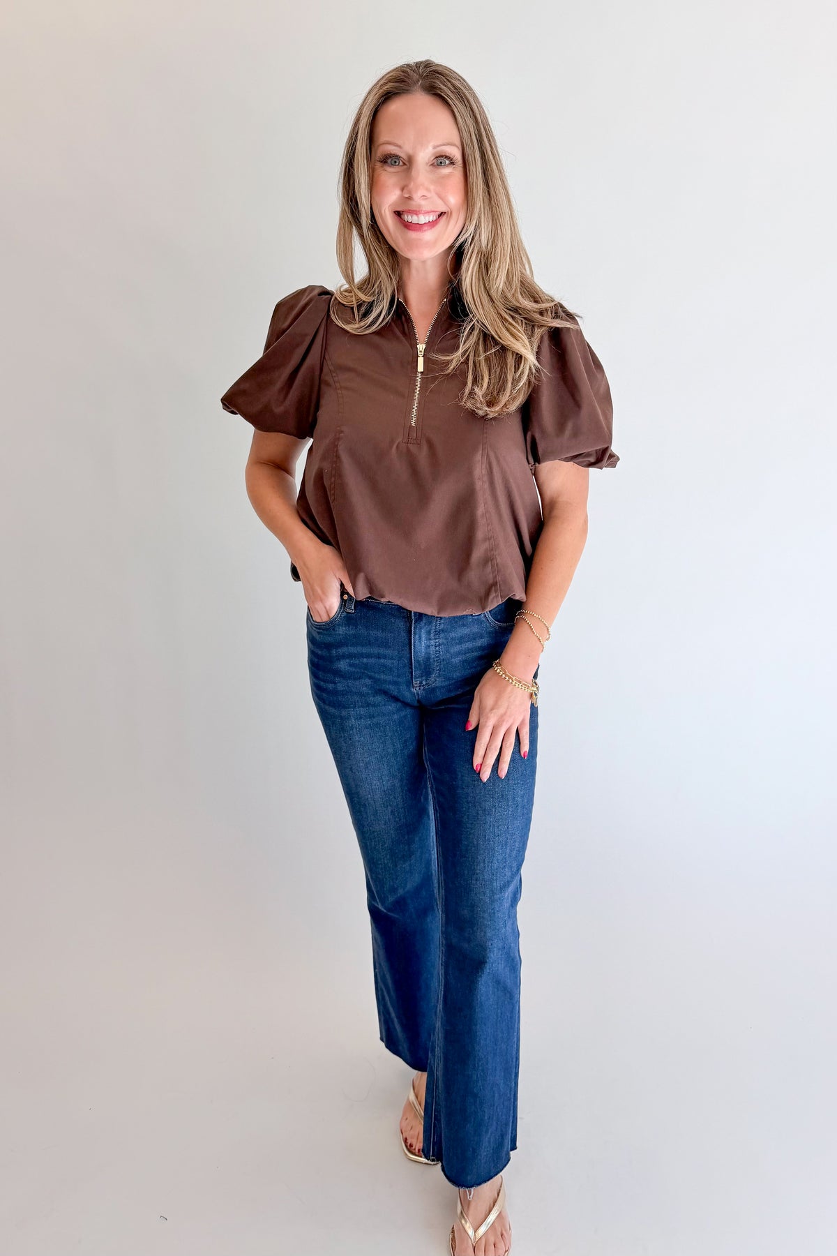 Front view of woman wearing Swagger Boutique’s Brown Zippered Bubble Hem Top by Entro with jeans against white studio background.