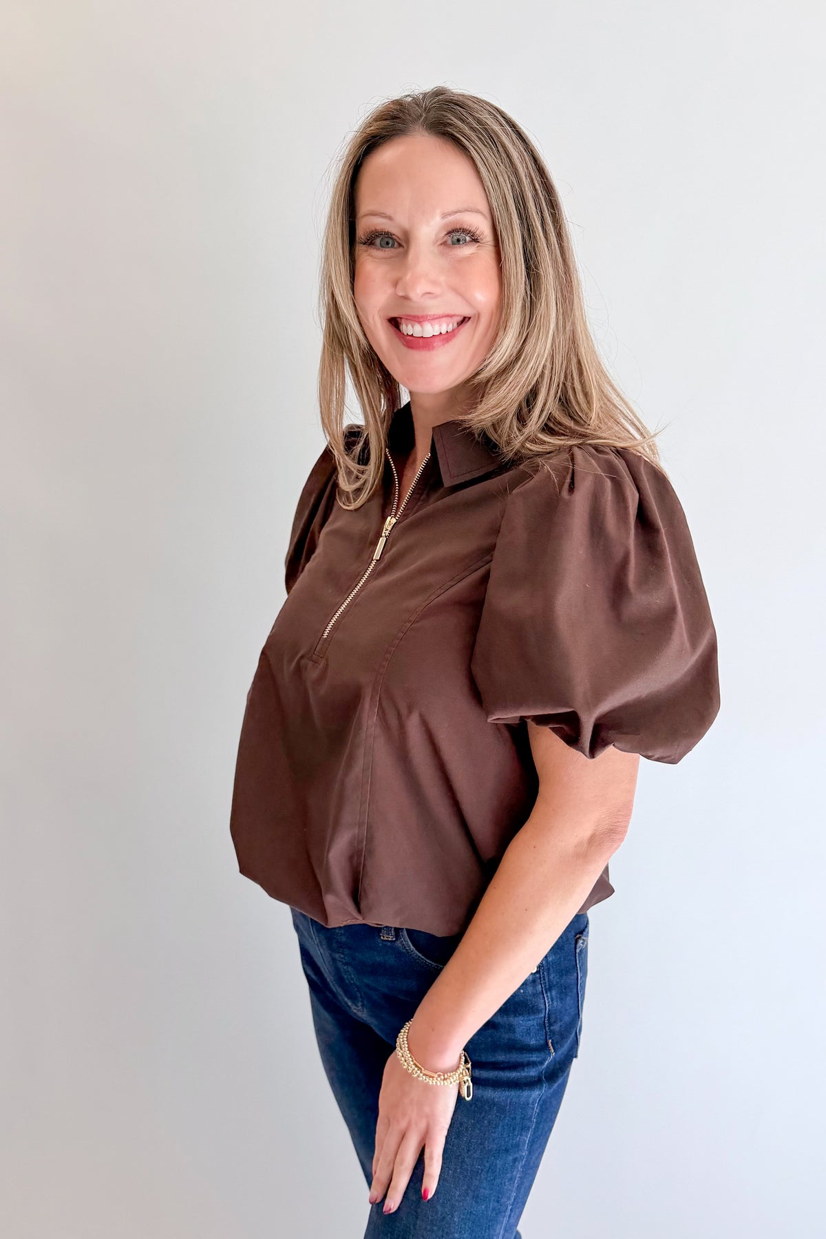 Side view of woman wearing Swagger Boutique’s Brown Zippered Bubble Hem Top by Entro with jeans against white studio background.