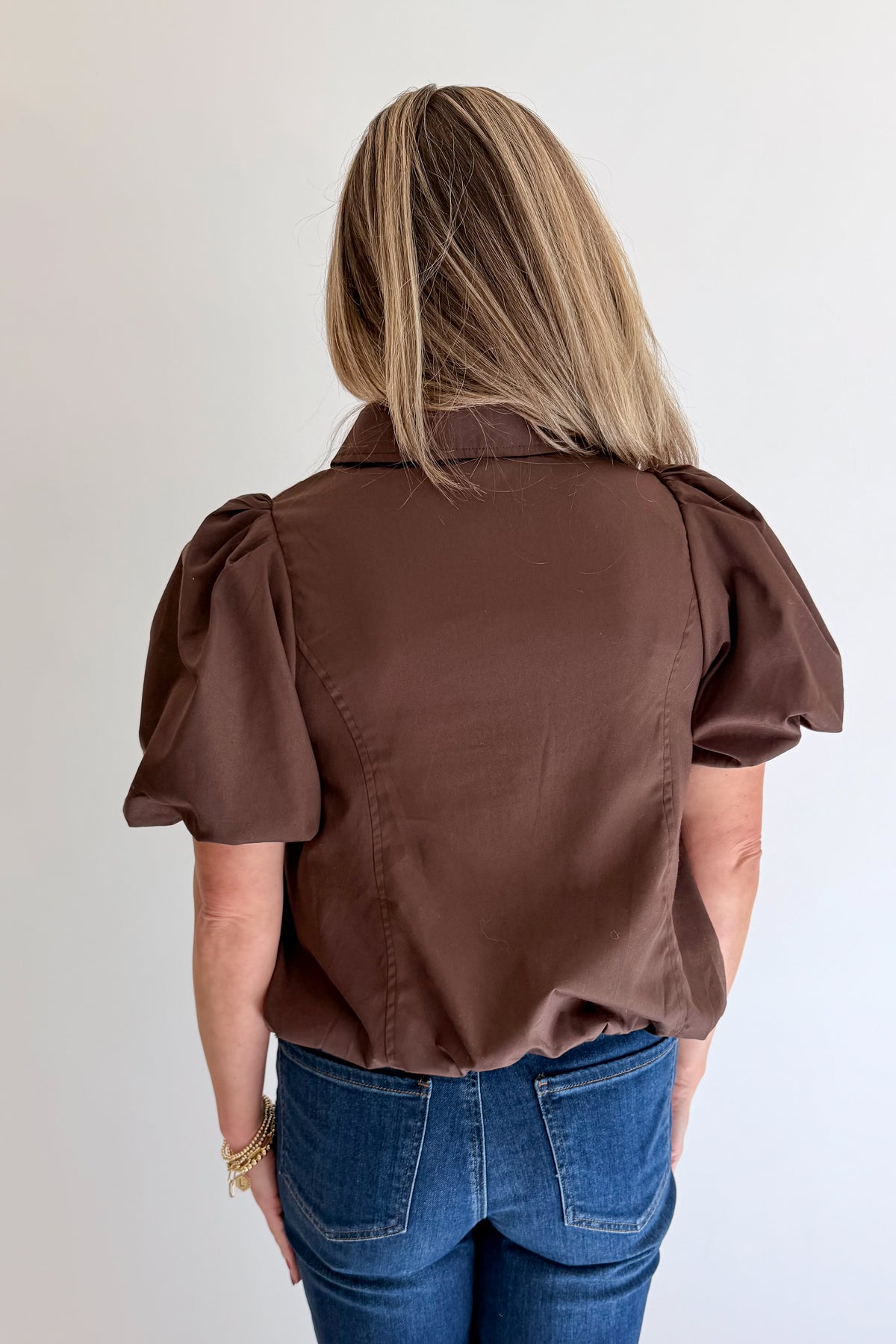 Back view of woman wearing Swagger Boutique’s Brown Zippered Bubble Hem Top by Entro with jeans against white studio background.